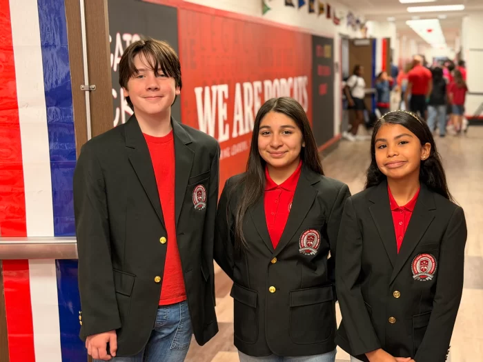 Three junior high or elementary students wearing school blazers and smiling in a school hallway with a "We Are Wildcats" sign. (Pathways: General School Choice, Magnet Program)