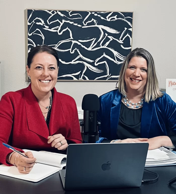 Podcast co-hosts Shereen El-Domeiri (right, blue blazer) and Nicola Hobeiche (left, red blazer) smiling while sitting at a desk with a microphone and laptop, recording an episode of Counsel Brew. 