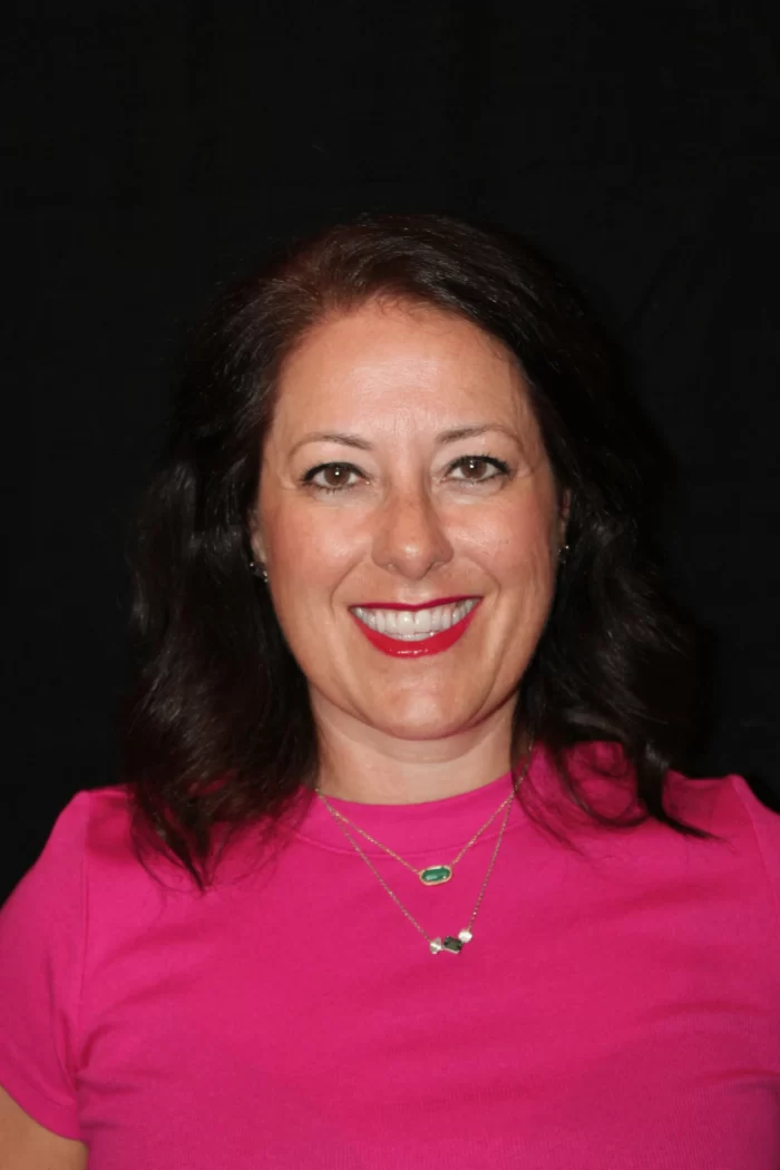 Professional headshot of Nicola Hobeiche, co-host of Counsel Brew, smiling with bright lipstick and wearing a pink top.