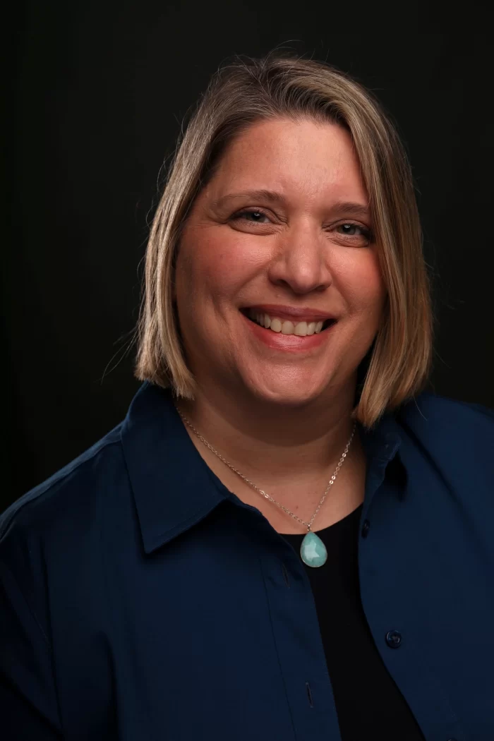 Podcast co-hosts Shereen El-Domeiri (right, blue blazer) and Nicola Hobeiche (left, red blazer) smiling while sitting at a desk with a microphone and laptop, recording an episode of Counsel Brew.
