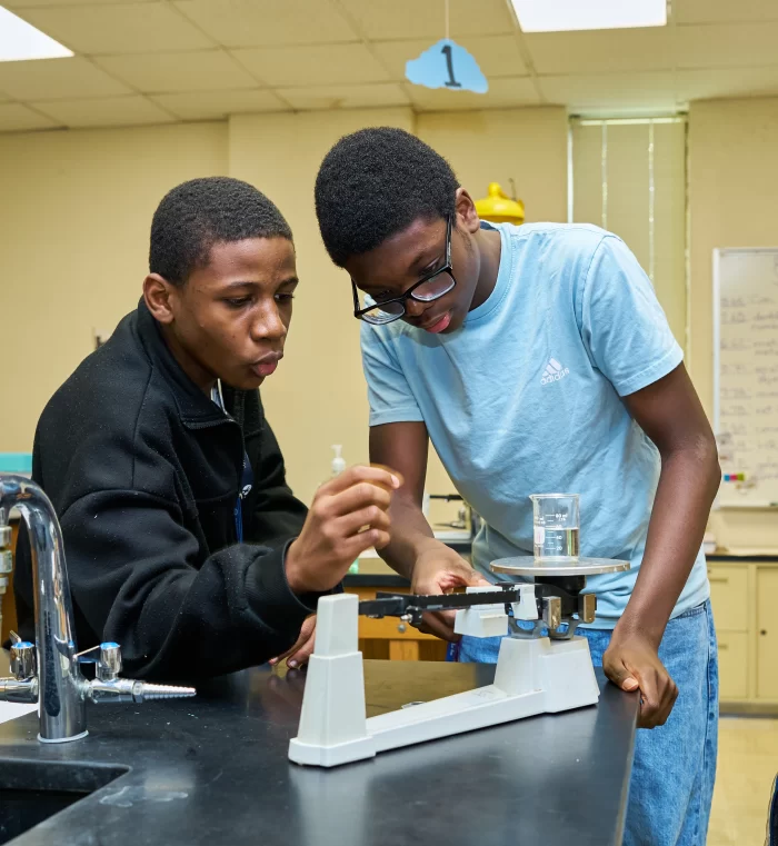 Two students conducting a chemistry experiment with a triple beam balance and beaker in a Richardson ISD science classroom.