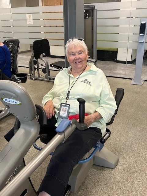 An older woman smiling while exercising on a NuStep recumbent cross trainer in the Richardson Senior Center fitness area.