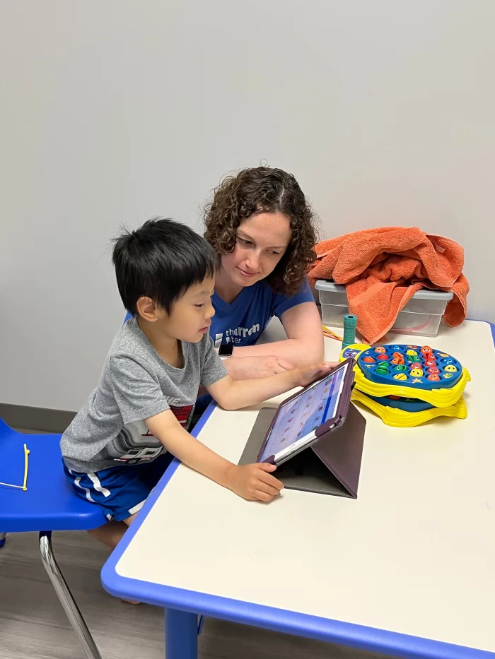 A therapist and a young boy working together on a tablet during a learning session at a desk.