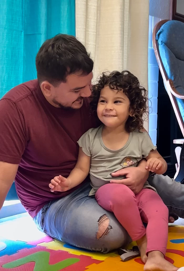 A father smiling and holding his young daughter during a home-based therapy session.