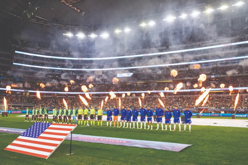 A wide view of a packed soccer stadium during a national anthem ceremony. The USA and Mexico teams stand in lines on the pitch, flanked by massive pyrotechnic flames and smoke, with a large American flag in the foreground.