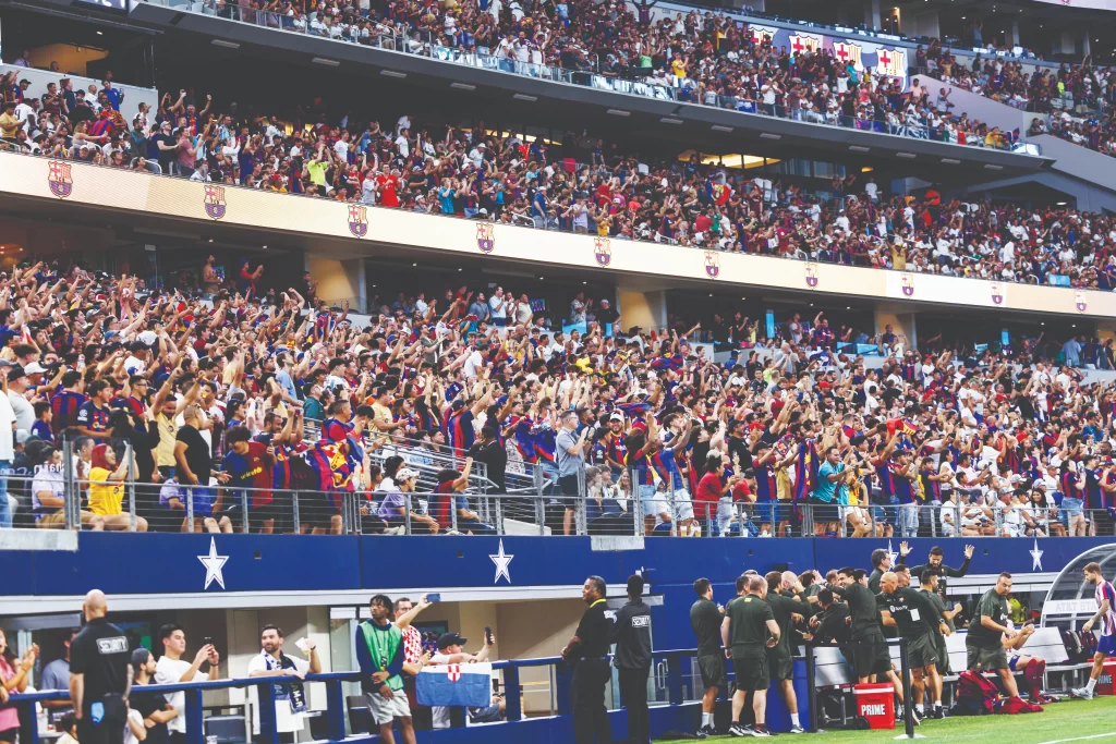 A multi-level stadium stands filled to capacity with cheering fans wearing various soccer jerseys. Many have their arms raised in excitement during a match.