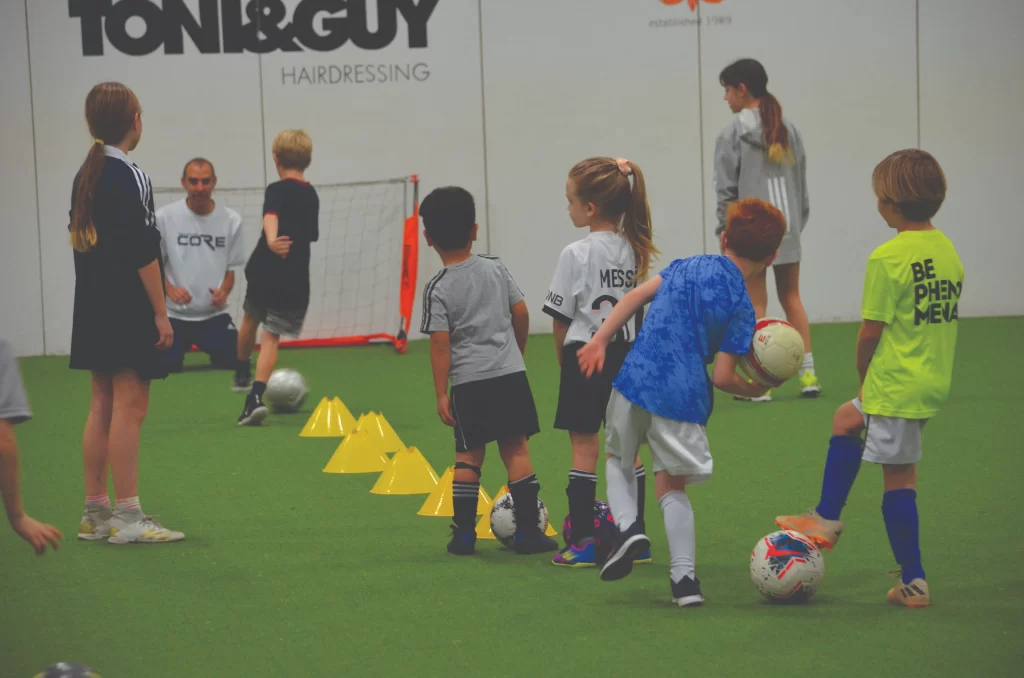 A group of young children participate in a soccer training session on an indoor green turf. They are lined up behind yellow cones, practicing ball control in front of a small goal.