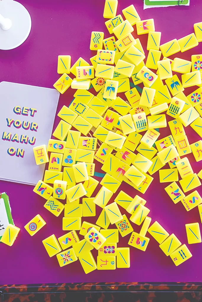 A scattered pile of vibrant yellow mahjong tiles with colorful modern engravings on a purple surface. A card next to the tiles reads "GET YOUR MAHJ ON"