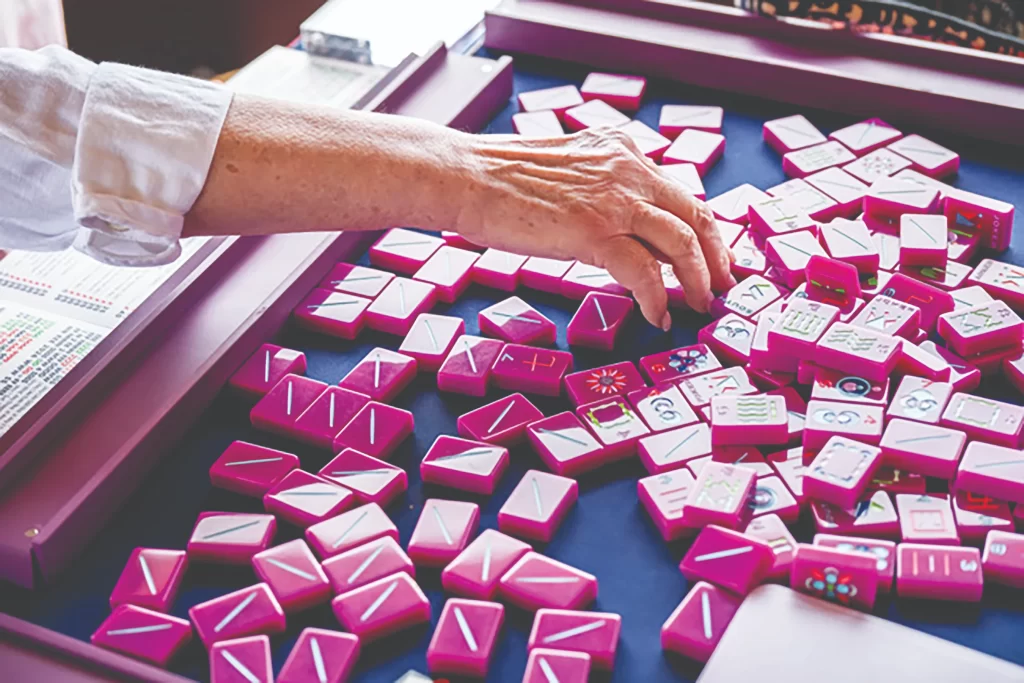 A close-up shot of a hand reaching for a pink mahjong tile among a large spread of tiles on a blue felt-lined game tray.