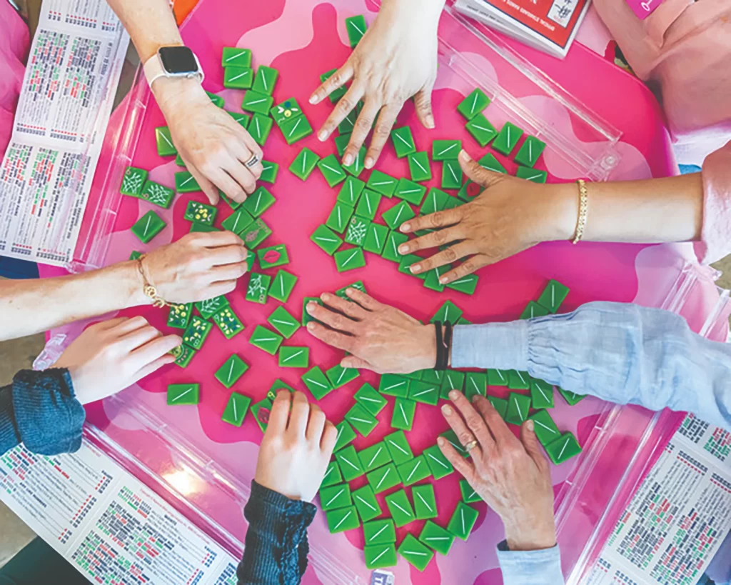 A top-down view of six hands reaching toward a large pile of bright green mahjong tiles on a pink tabletop. Official NMJL scorecards are visible at the edges of the table.
