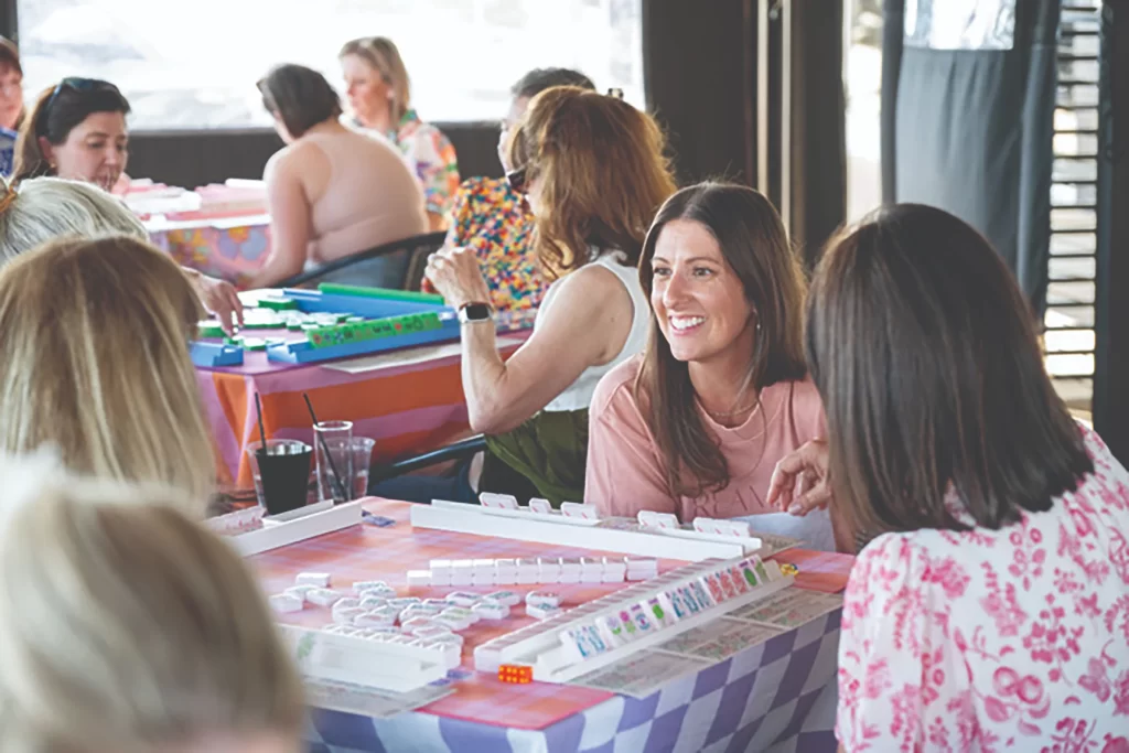 A woman with long brown hair smiles broadly while sitting at a mahjong table with friends. In the background, several other groups are playing at tables with colorful tablecloths.