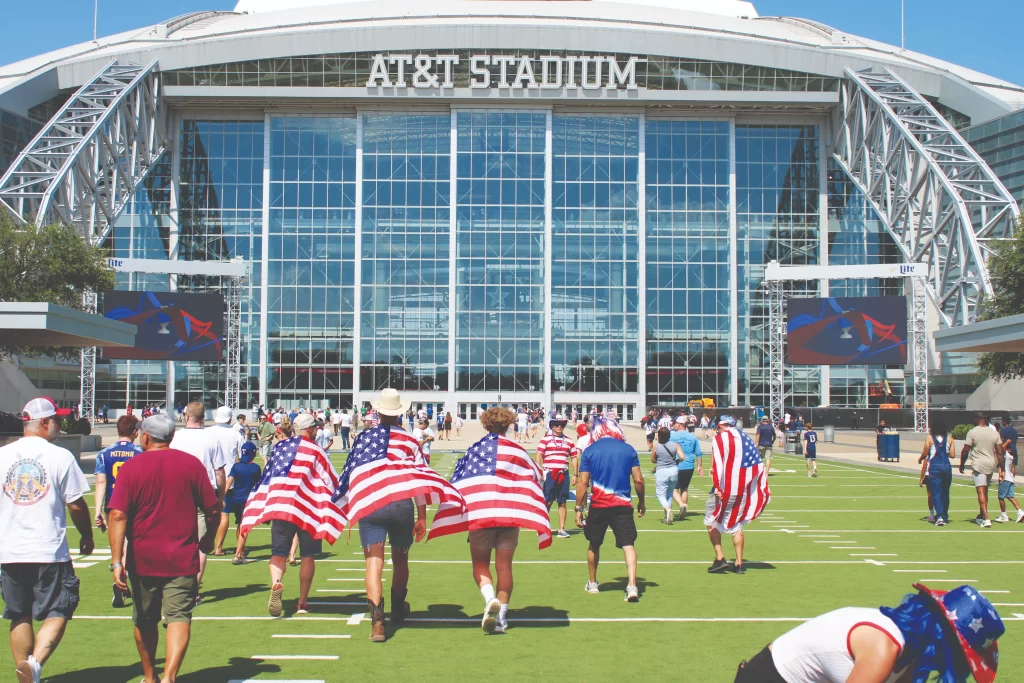 Several fans wearing American flags as capes walk across a green plaza toward the massive glass entrance of AT&T Stadium under a clear blue sky.