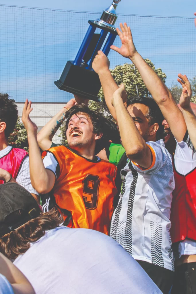 A young man in an orange scrimmage vest laughs with joy while holding a large blue and silver trophy high above his head, surrounded by cheering teammates.