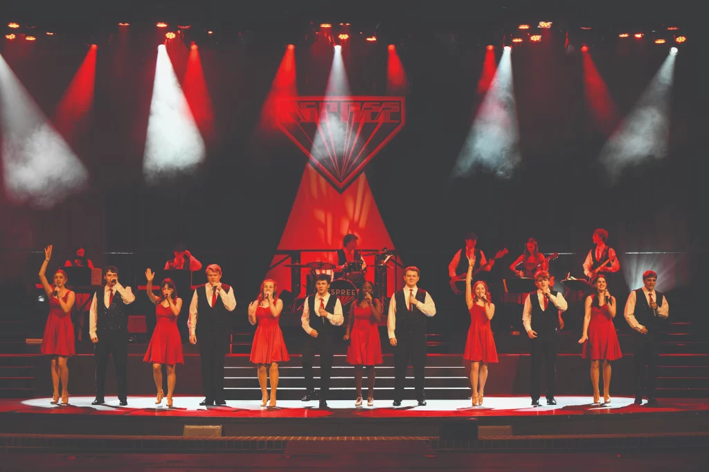 A group of young students in traditional Ballet Folklórico attire performing on an outdoor stage. The girls are wearing vibrant, multi-colored ruffled skirts and the boys are in traditional charro suits.