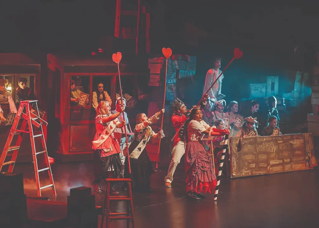 A close-up of a student in an art classroom, focused on sketching a detailed portrait in a sketchbook. Other art supplies and student works are visible in the background.