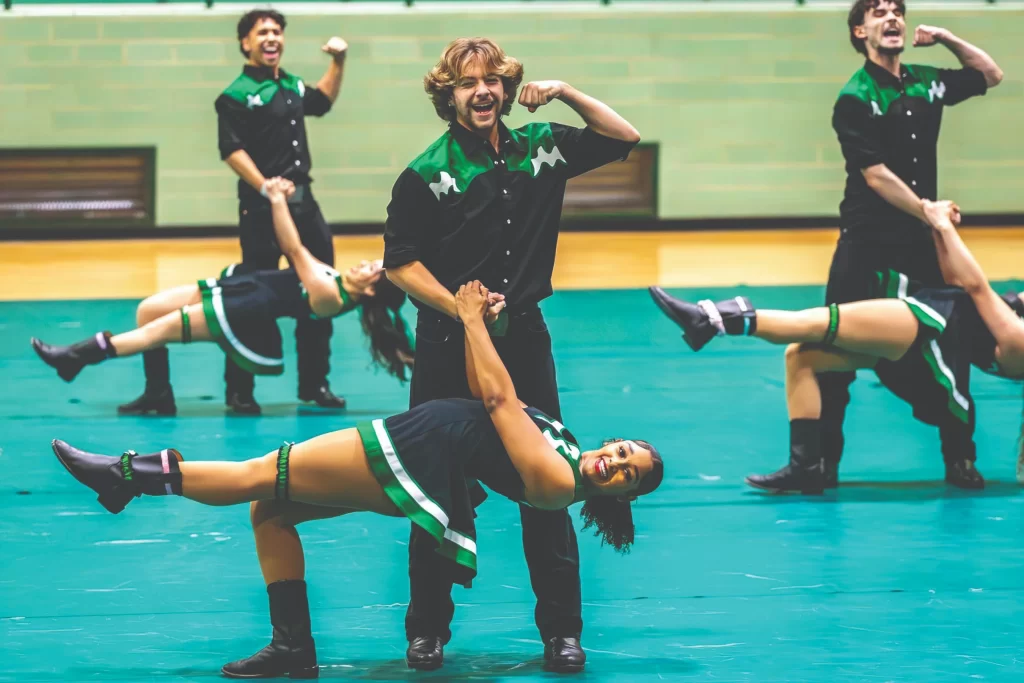 A student musician in a formal black tuxedo playing a violin during a school orchestra performance. The background shows other members of the string section in a concert hall setting.