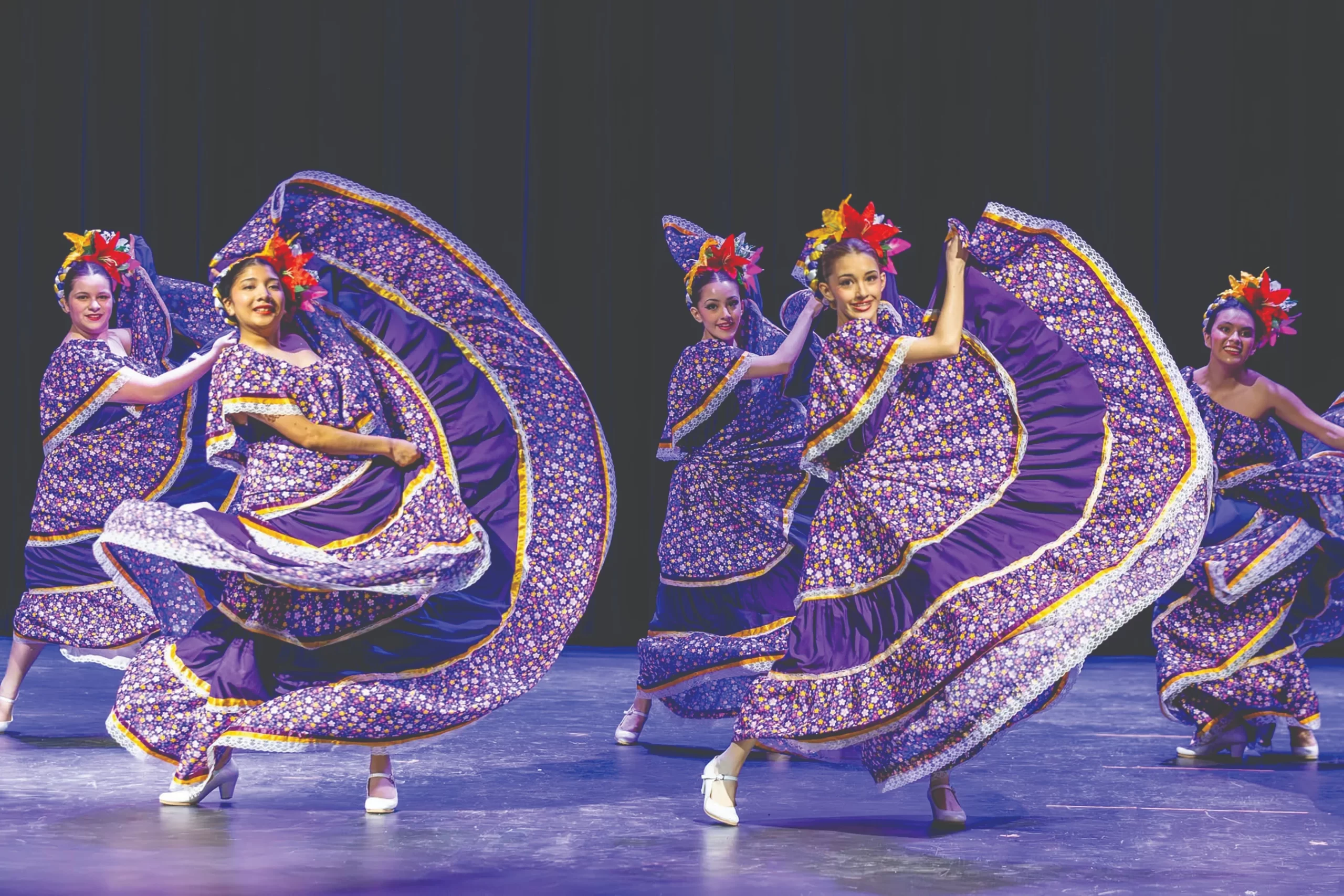 A group of elementary school students on stage during a musical production. They are in colorful costumes and captured mid-song with expressive faces and hand gestures.