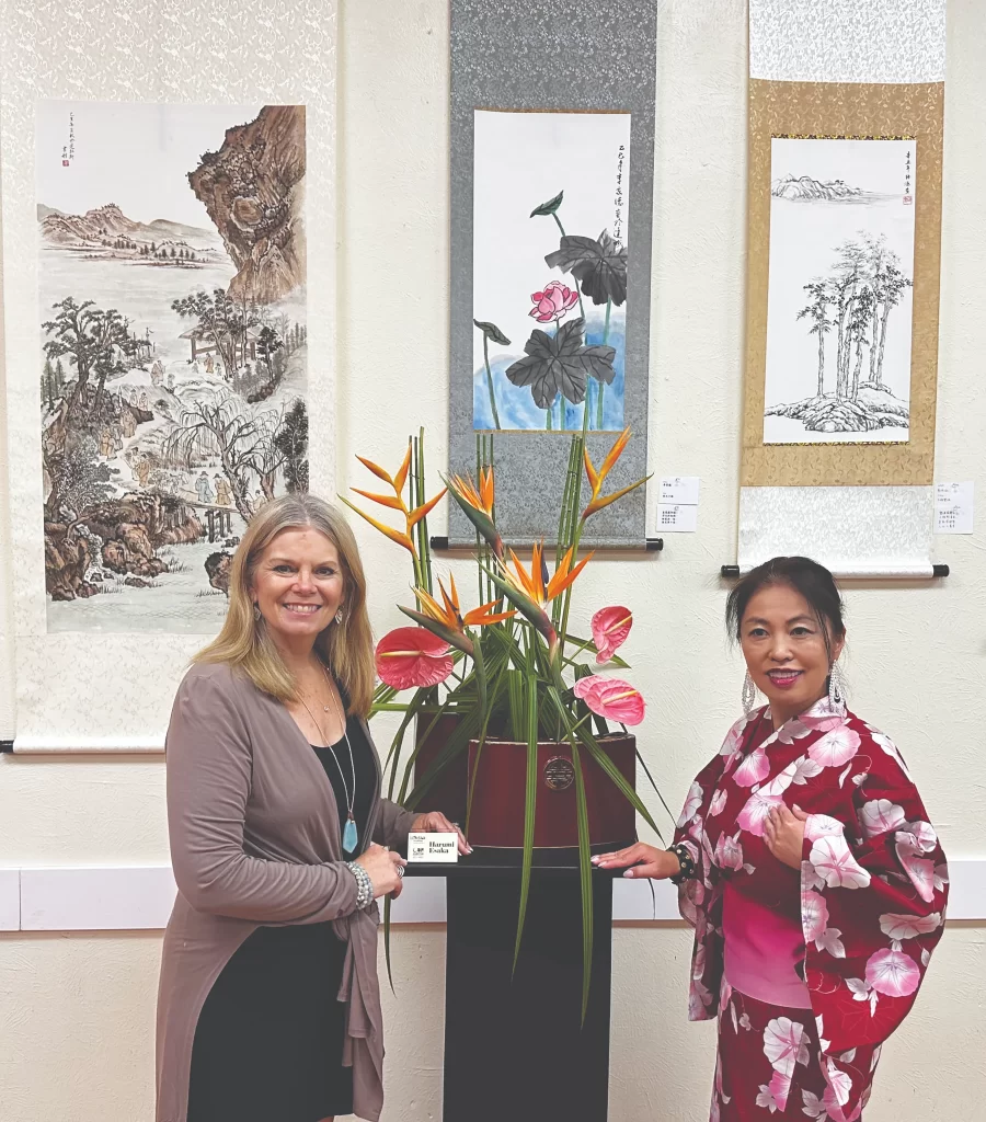 A professional portrait of Sandy Palisch smiling, wearing a teal blazer and a layered necklace. She is standing in a brightly lit, modern indoor space.
