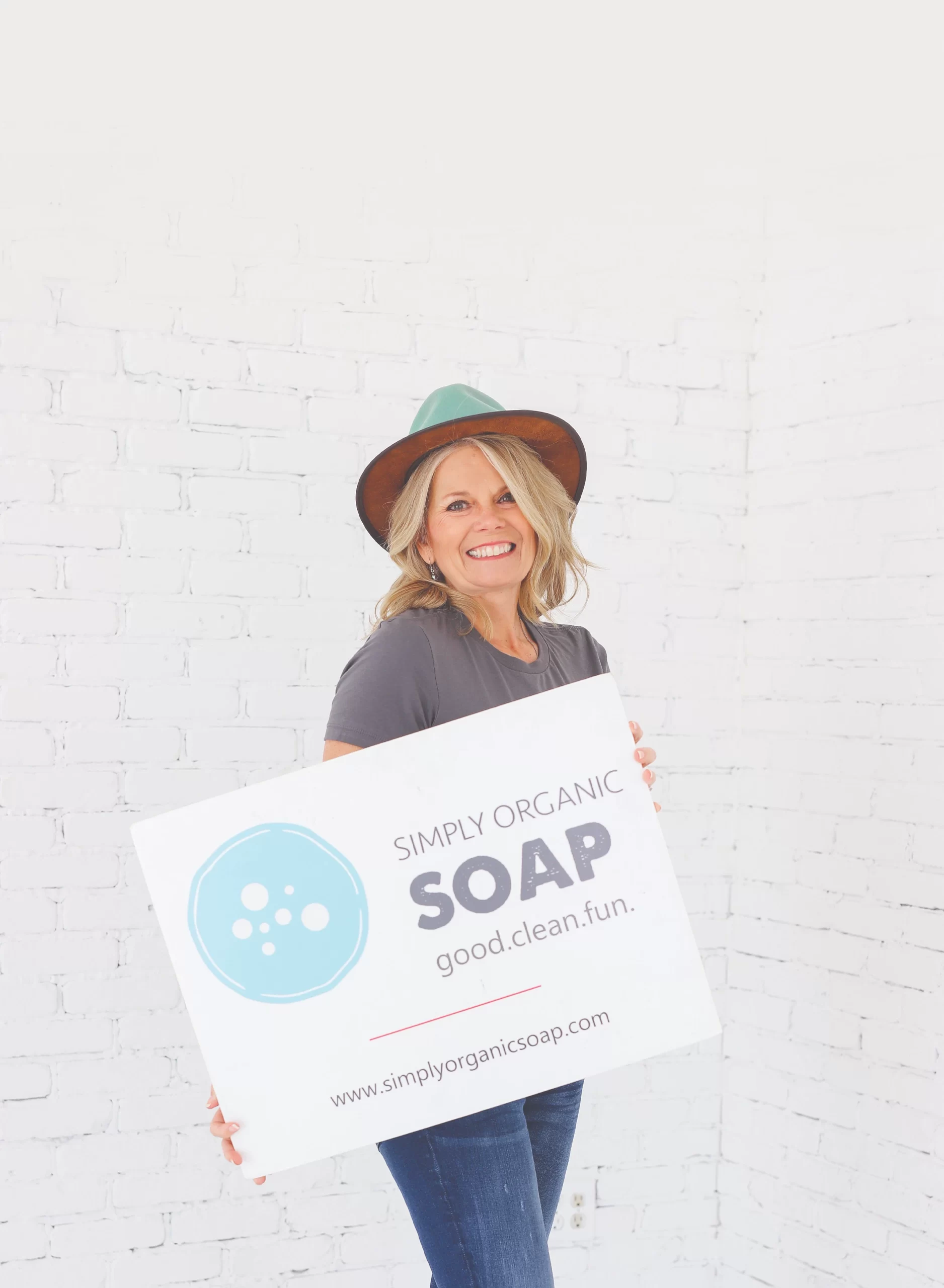Sandy Palisch smiling warmly while standing in front of a shelf filled with "Simply Organic Soap" products. She is wearing a casual denim shirt.