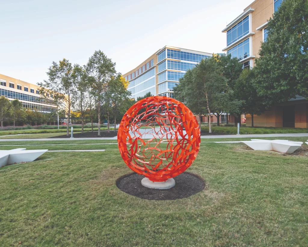 A large-scale public art installation in a paved plaza featuring two tall, sleek metallic sculptures that curve toward each other against a blue sky.