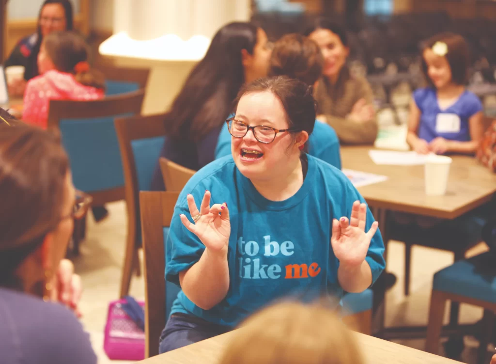Close-up of a "To Be Like Me" LEADer wearing glasses and a blue organization t-shirt. She is smiling and gesturing with her hands while engaging in a conversation with someone off-camera at a table.