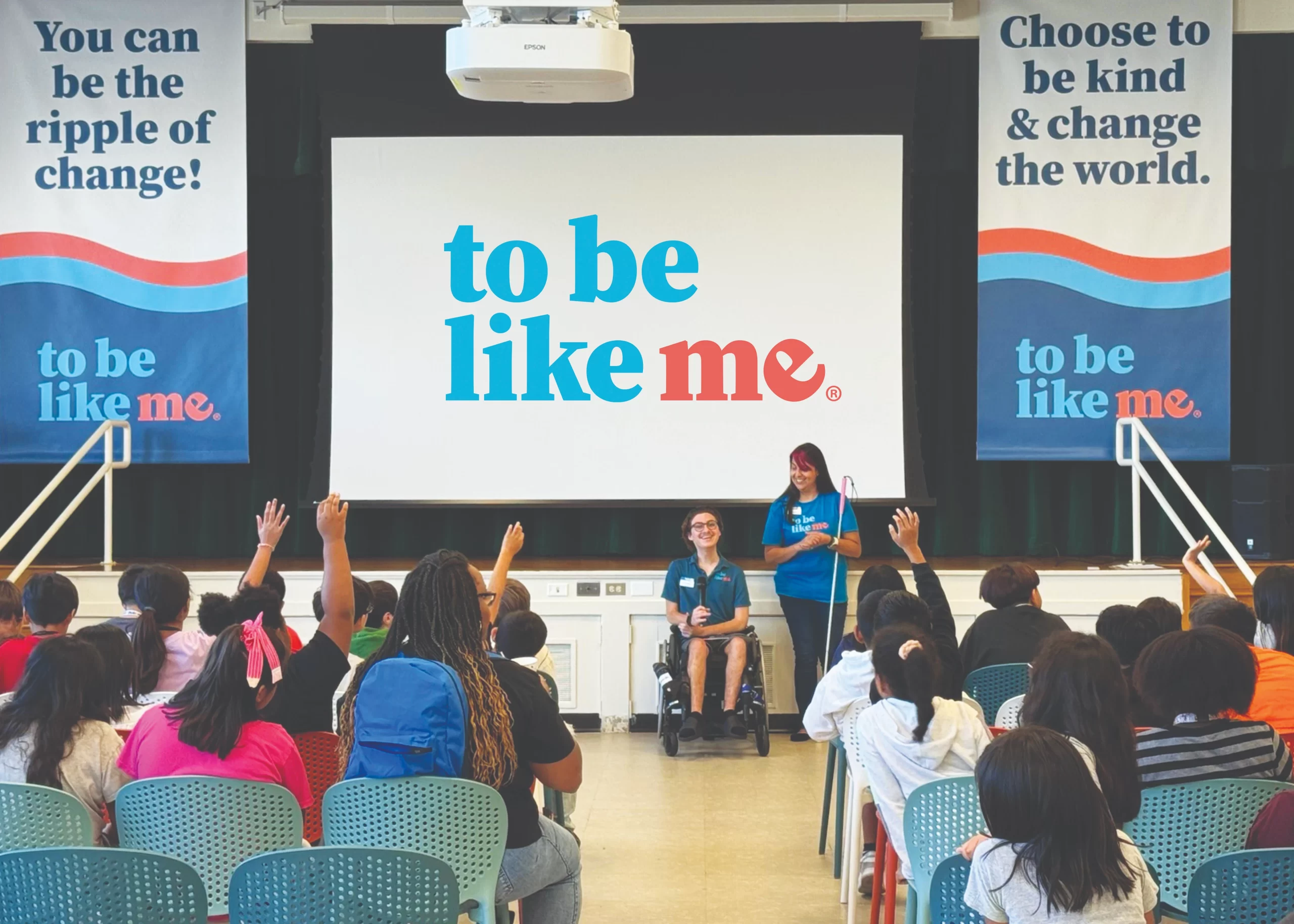 A "To Be Like Me" presentation in a school auditorium. Two LEADers—one in a wheelchair and one standing with a white cane—speak to a large group of seated students who have their hands raised.