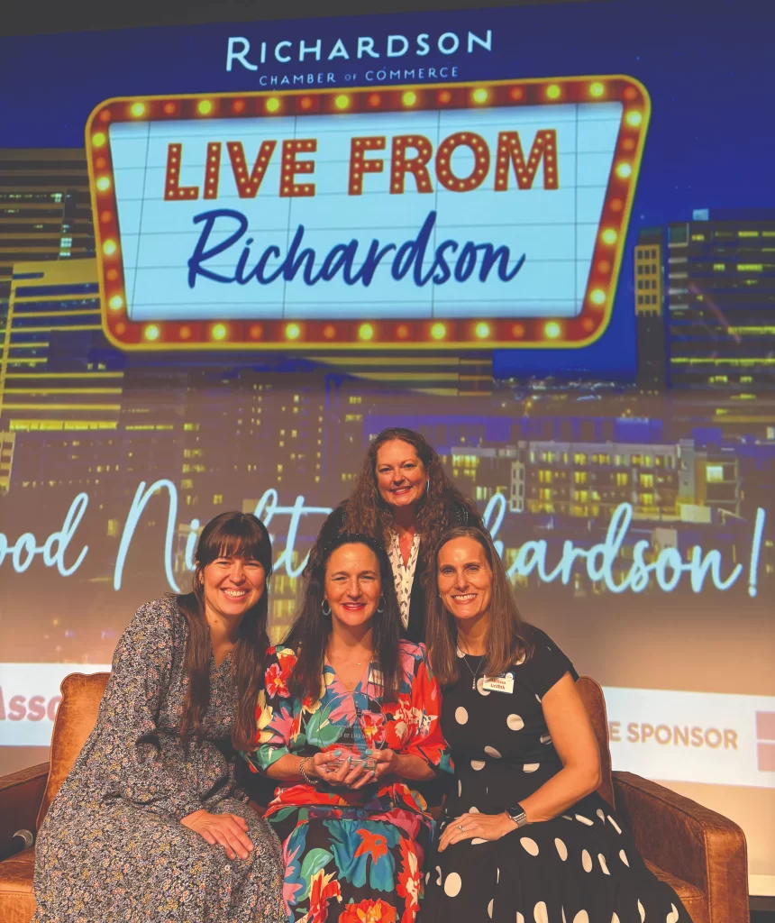 Four women smile while sitting on a couch at an event. The woman in the center holds a glass award. The background screen reads "Richardson Chamber of Commerce: Live from Richardson!"