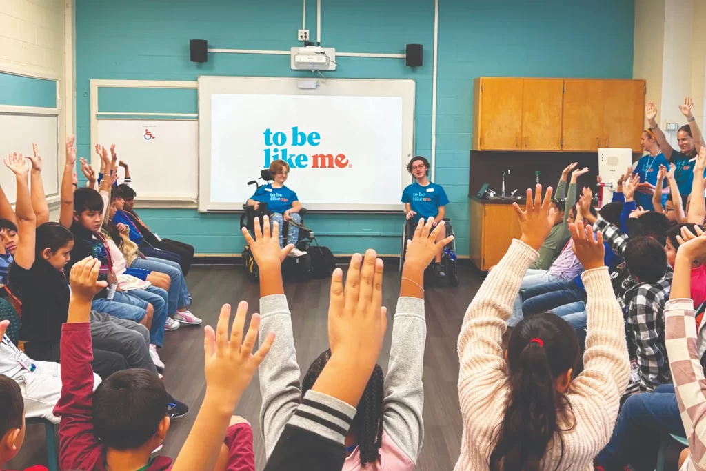Two "To Be Like Me" LEADers in wheelchairs sit at the front of a colorful classroom. A semi-circle of elementary students face them with their hands raised in an interactive Q&A session.