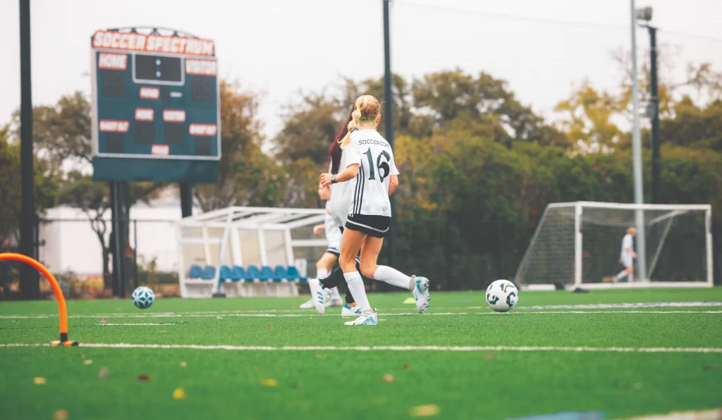 A young girl in a white soccer jersey with the number 16 runs across an outdoor turf field during a bright day. A scoreboard in the background displays "Soccer Spectrum."