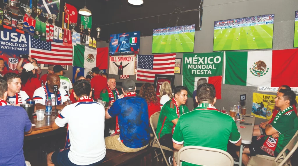 A crowded indoor watch party at Soccer Spectrum. Fans wearing USA and Mexico jerseys sit at tables with drinks, surrounded by flags and multiple TV screens showing a soccer match.