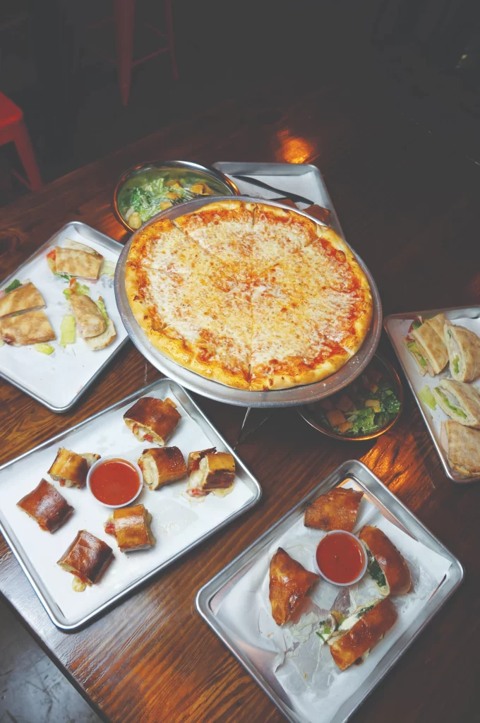 An overhead view of a wooden dining table spread at Azzurri Pizzeria + Bar featuring a large cheese pizza, sliced paninis, dynamic side salads, and dipping rolls.