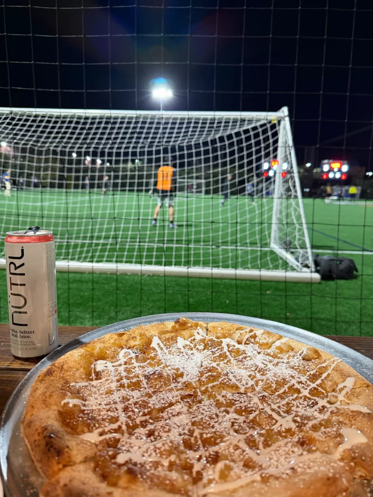 A fresh, specialty dessert pizza dusted with powdered sugar sitting on a table alongside a can of NÜTRL seltzer, with an indoor soccer goal and league game happening in the background.