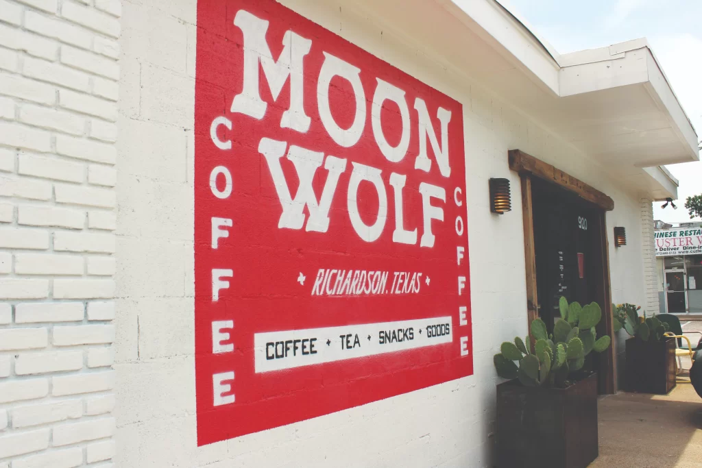 A bold red and white painted mural on a white brick building facade reading "Moon Wolf Coffee, Richardson, Texas" near a storefront entrance with potted cacti.