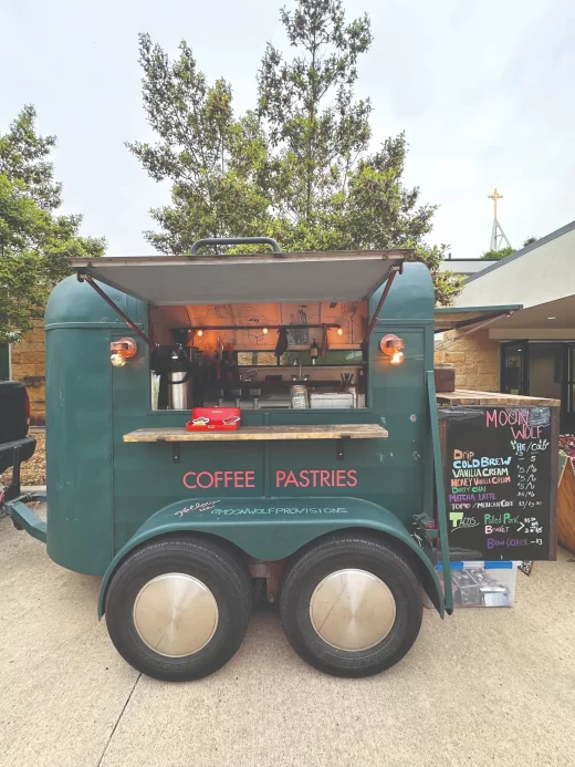 A retro green goat trailer converted into a mobile coffee shop booth, featuring wood counters, hanging light bulbs, and a handwritten chalkboard menu.