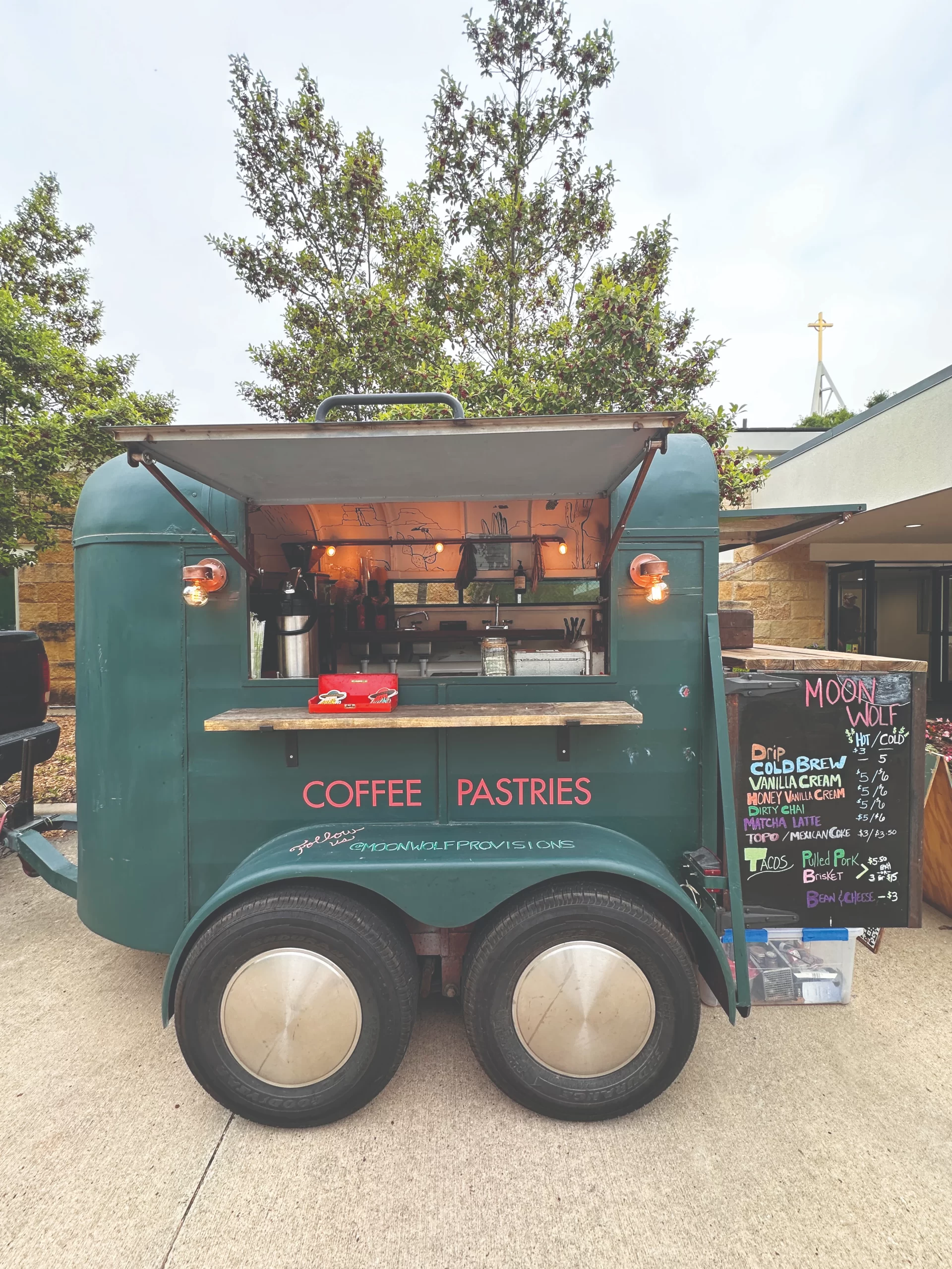 A retro green goat trailer converted into a mobile coffee shop booth, featuring wood counters, hanging light bulbs, and a handwritten chalkboard menu.
