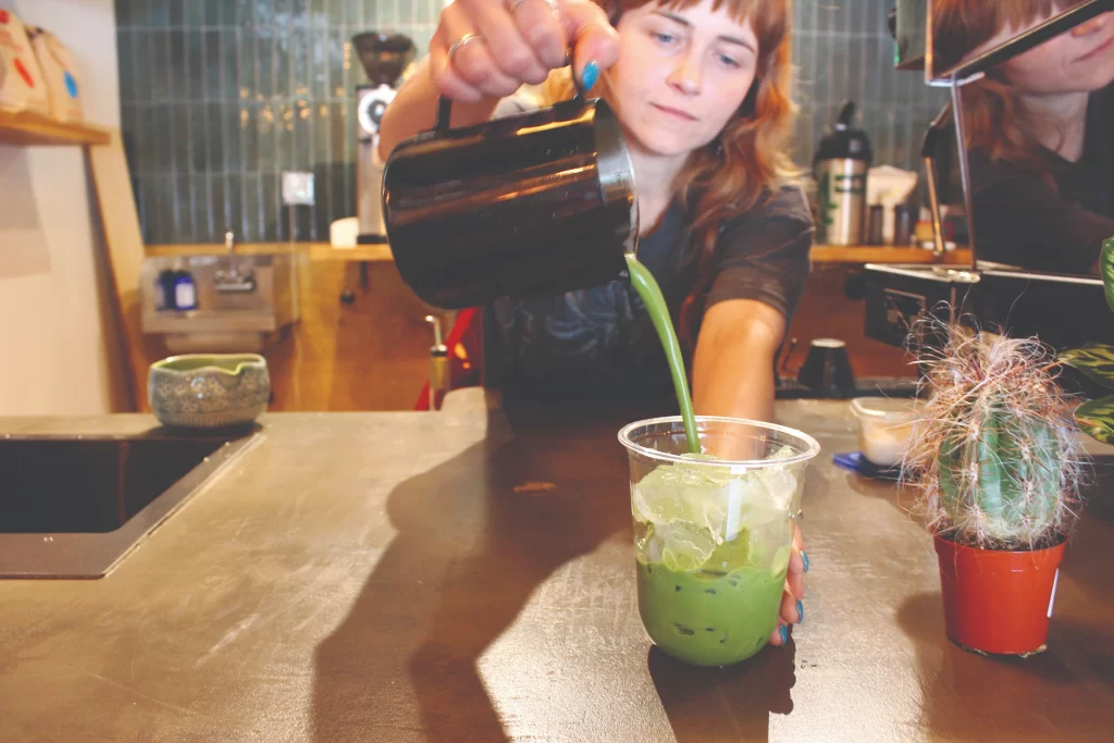 A barista carefully pouring bright green ceremonial-grade iced matcha from a black pitcher into a clear plastic cup on a concrete counter next to a tiny potted cactus.