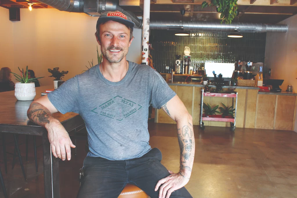 Owner Richie Conry wearing a gray t-shirt and a baseball cap, smiling while sitting at a custom wooden bar inside his hand-built coffee shop.
