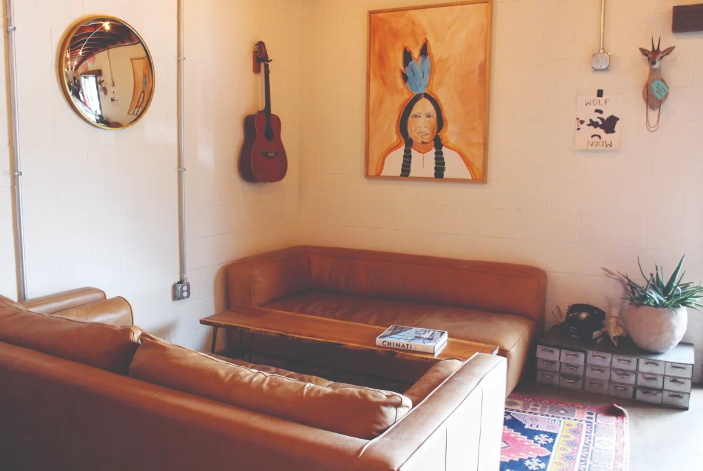 A cozy coffee shop seating corner featuring tan leather couches, a live-edge wooden coffee table, an patterned rug, acoustic guitar, and unique wall art.