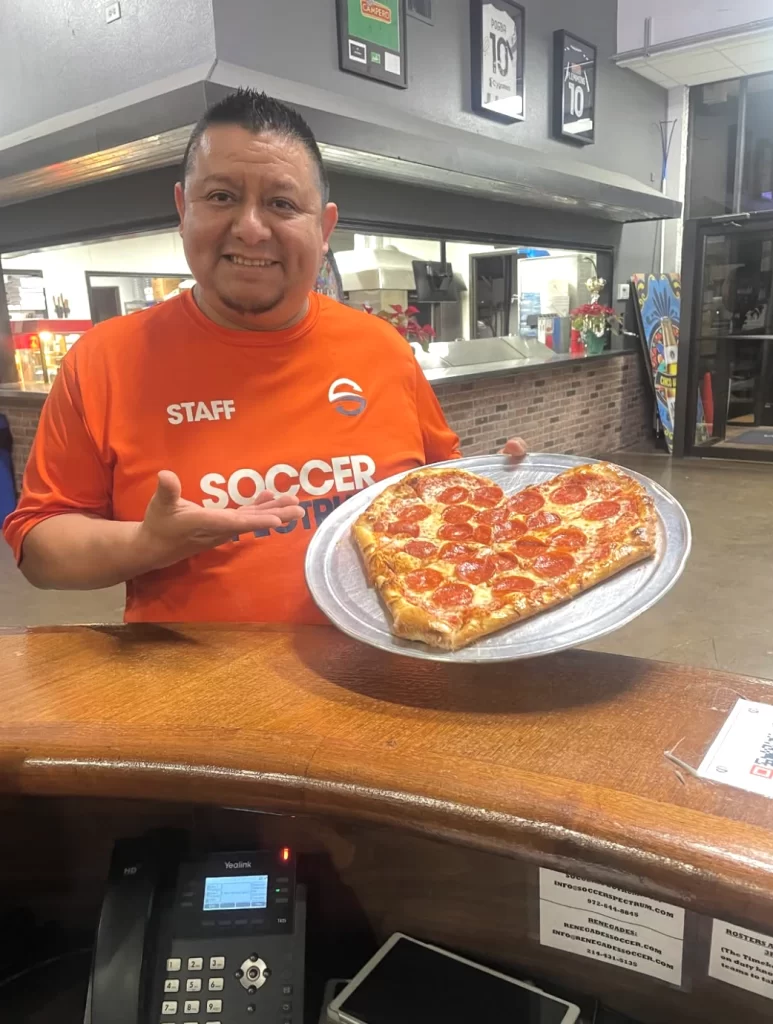 Head Chef Eloy Tejeda Costeno standing proudly at the service counter in an orange Soccer Spectrum shirt, presenting a fresh, heart-shaped pepperoni pizza.