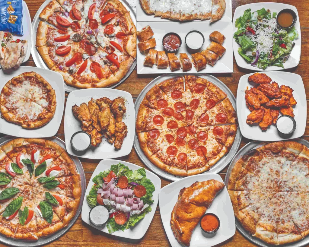 A massive overhead feast of Italian comfort food, including multiple specialty pizzas, chicken wings, fresh calzones, side salads, and garlic rolls.