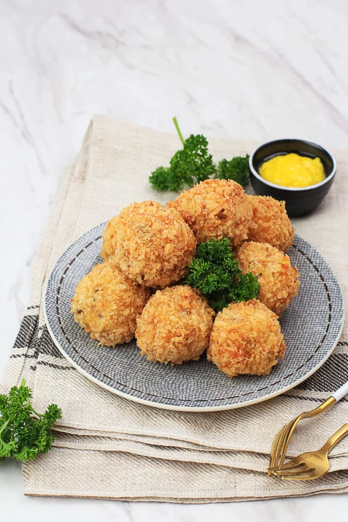 A plate of golden, crispy bitterballen garnished with fresh parsley, served on a grey patterned dish next to a small bowl of yellow mustard.
