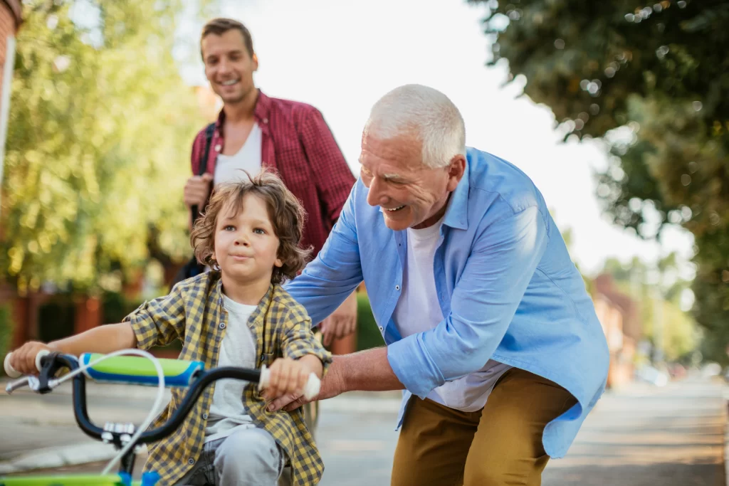 A joyful grandfather helping his young grandson steady a bicycle on a neighborhood street while the middle-aged father smiles in the background.