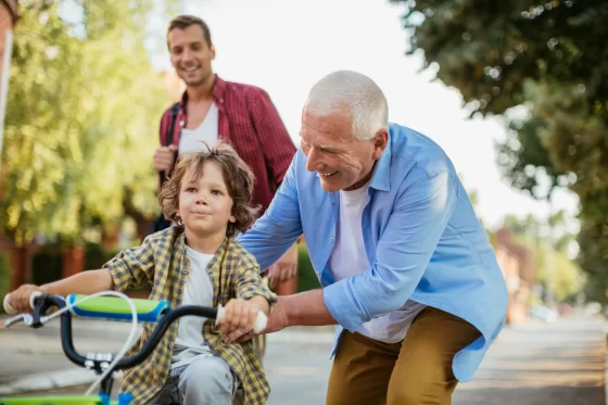 A joyful grandfather helping his young grandson steady a bicycle on a neighborhood street while the middle-aged father smiles in the background.