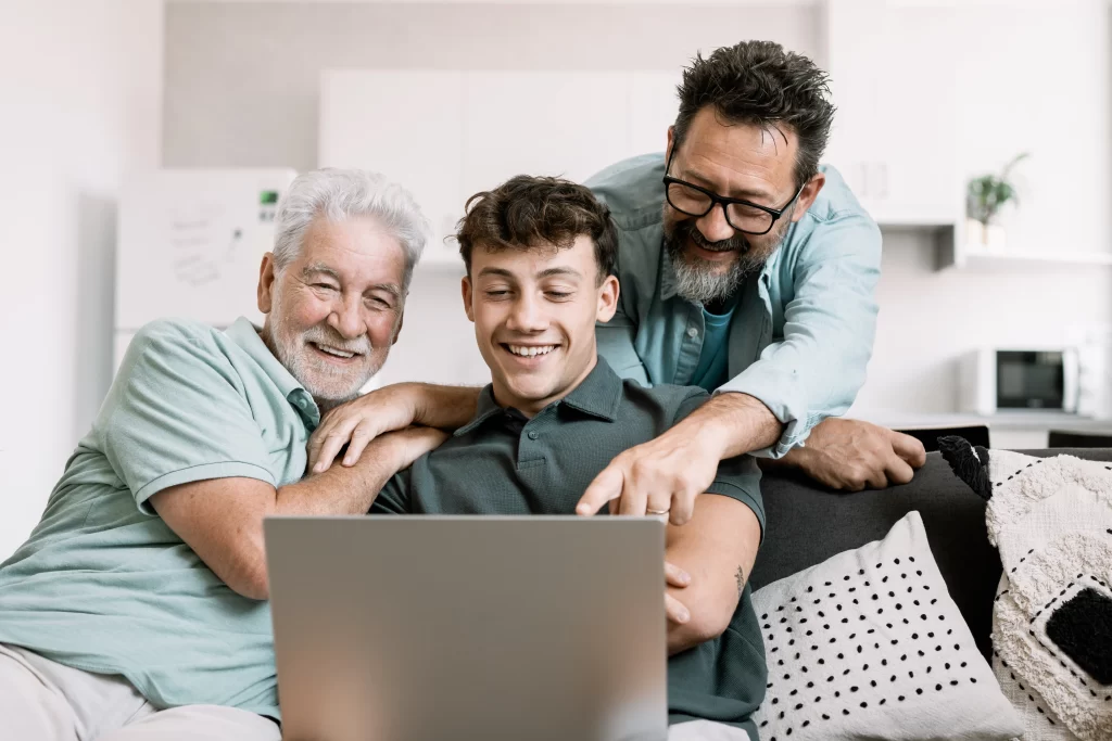 Three generations of men sitting on a couch looking at a laptop screen—a smiling grandfather, a young adult grandson, and a middle-aged father pointing at something on the screen.