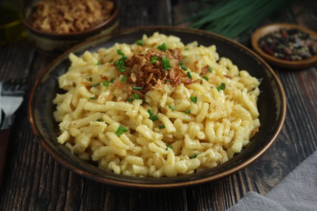A close-up of a warm bowl of Käsespätzle, featuring soft German egg noodles tossed in melted cheese and topped with crispy fried onions and fresh chives.