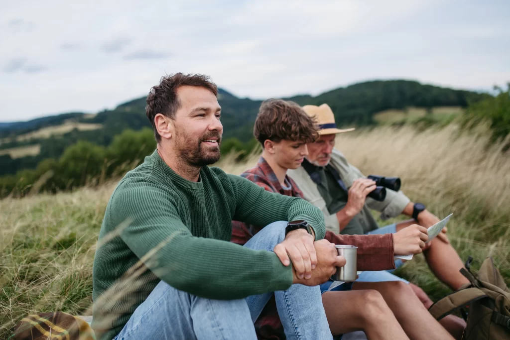 A middle-aged man in a green sweater sitting on a grassy hill with his teenage son and elderly father, who holds a map and binoculars.