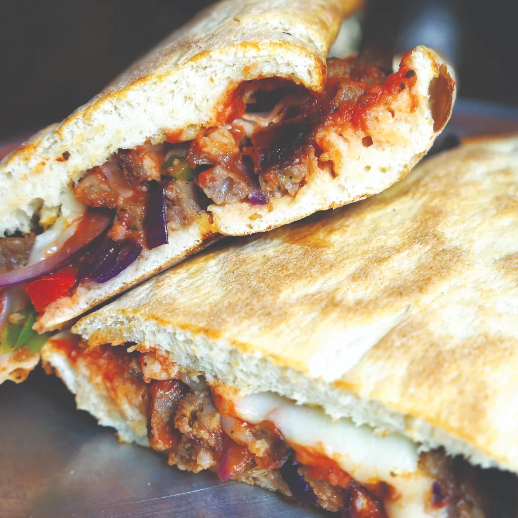 A close-up shot of a sliced panini sandwich at Azzurri Pizzeria + Bar, showcasing melted cheese, meats, red onions, and fresh peppers inside a toasted, scratch-made crust.