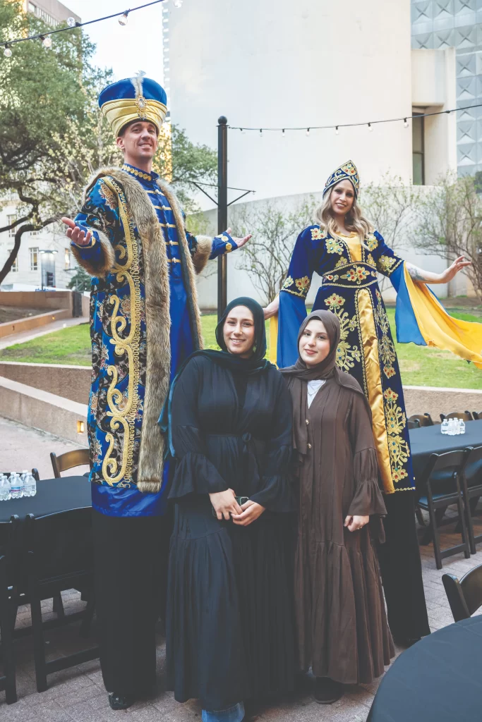Participants wearing ornate blue and gold traditional costumes pose with attendees at an outdoor community gathering in Richardson, Texas.