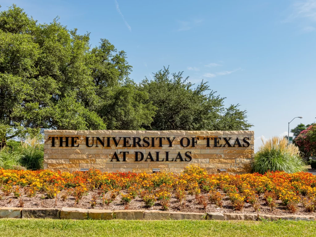 The University of Texas at Dallas entrance sign surrounded by landscaped flowers and trees in Richardson, Texas.